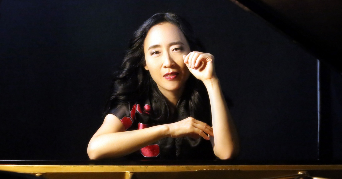 Woman sitting at a grand piano, looking forward with hand raised near her face.