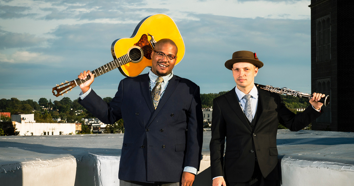 Two men in suits stand outside; one holds a guitar, the other holds a clarinet.
