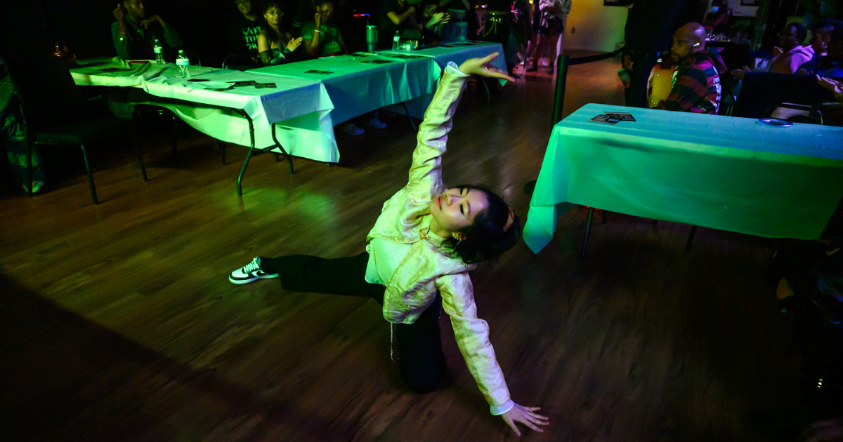 A person poses on a wooden floor in front of tables at an indoor event.