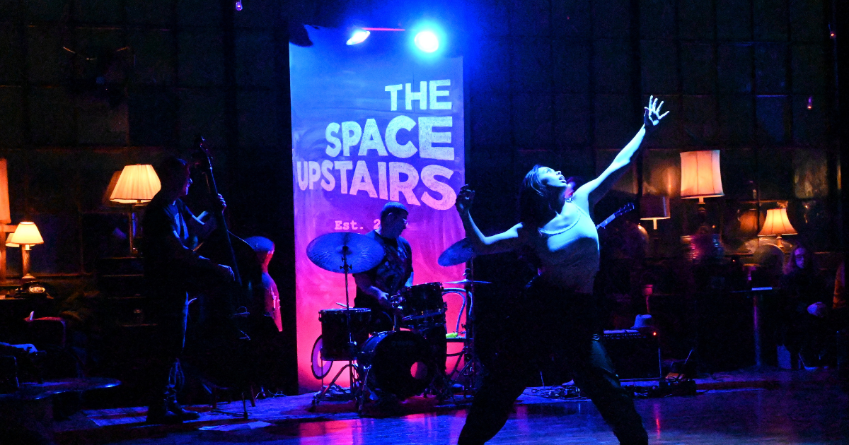 A dancer performs on stage with a band under blue and purple lighting.