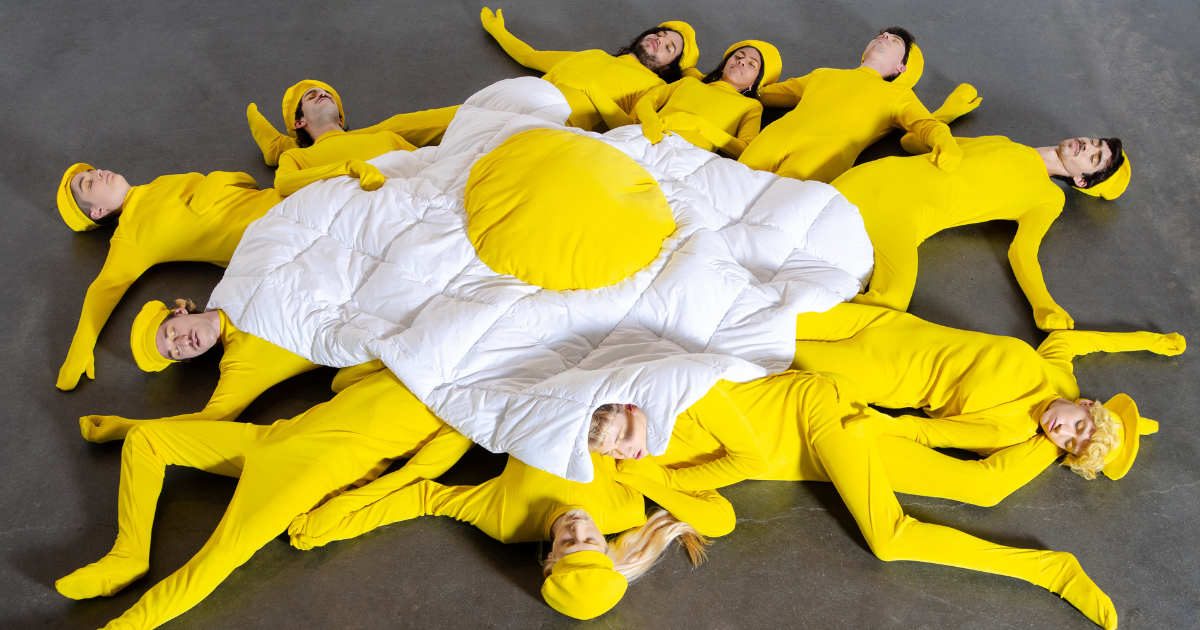 People in yellow bodysuits form the shape of a fried egg on a gray floor.