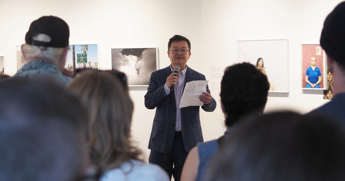 Someone speaking into a microphone at a gallery event while a crowd listens.