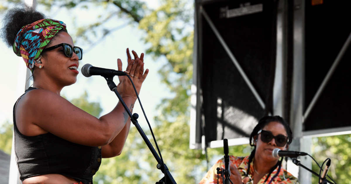 Two women perform on an outdoor stage, one singing and clapping, one playing guitar.
