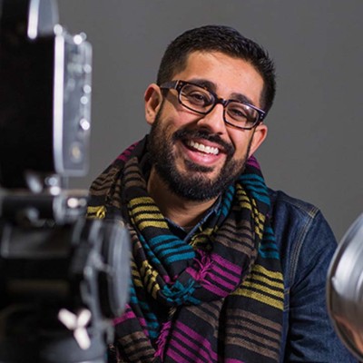 An image of Adil Mansoor, a theatre director and educator centering the stories of queer folks and people of color. He has brown skin and dark hair, a beard, and black glasses. He sits in front of a movie camera and light, wearing a striped, colorful scarf.