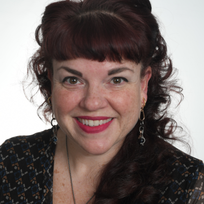 Smiling woman with dark auburn hair styled in a high curled updo, wearing red lipstick, dangling earrings, and a dark patterned top