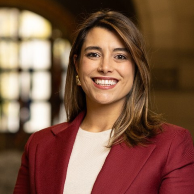 Woman in a maroon blazer smiling, standing indoors with blurred background.