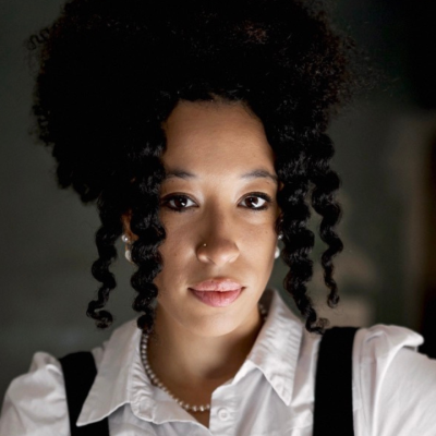 Portrait of a person with curly dark hair, wearing a white button-up shirt and dark vest