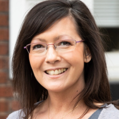 Smiling person with dark shoulder-length hair and rectangular purple-rimmed glasses, wearing a gray cardigan and a small heart-shaped pendant necklace