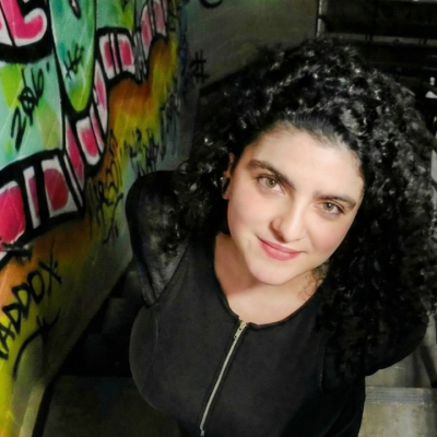 Woman with curly hair stands by graffiti-covered wall, looking up at the camera.