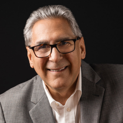Smiling man with short gray hair wearing black rectangular glasses and a gray blazer over a white button-up shirt