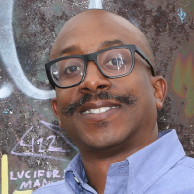 Smiling man with a dark mustache, black glasses, wearing a blue button-down shirt