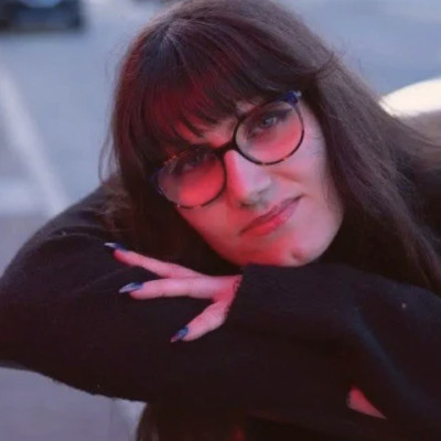 Woman with long straight dark hair and bangs, dark glasses, and a black long-sleeved shirt poses for a portrait on a city street