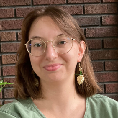 A smiling woman with shoulder-length brown hair wearing round glasses and grape-shaped earrings.