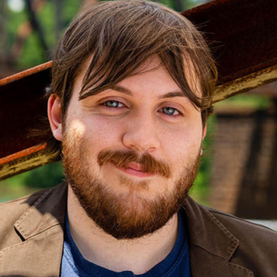 Close-up of a person with short brown hair and a full beard, wearing a dark jacket with a small name tag and a blue T-shirt