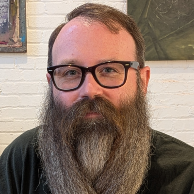 portrait of a person with dark-framed glasses and a long full beard, wearing a black T-shirt with white graphic text