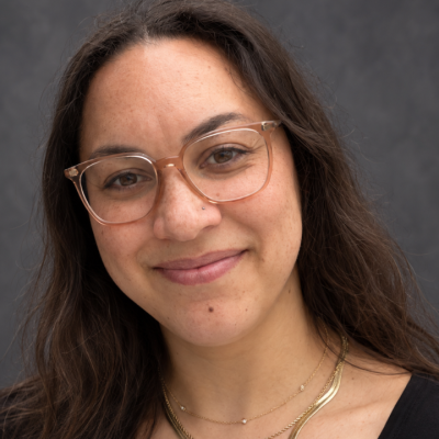Close-up studio portrait of a person with long dark hair, wearing clear-framed glasses, a layered gold necklace, and a black top