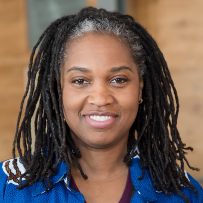 A smiling person with shoulder-length black locs wearing a bright blue jacket over a dark maroon top