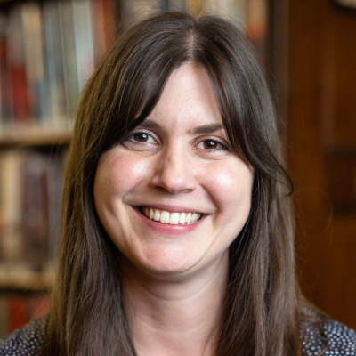 Smiling person with long dark brown hair, wearing a dark patterned top and a small gold necklace