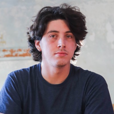 Latino person with short wavy dark hair, blue t-shirt, and a small tattoo on their arm