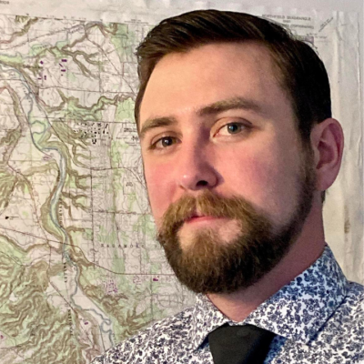 Portrait of a man with short dark hair and a full beard wearing a blue patterned shirt and black tie