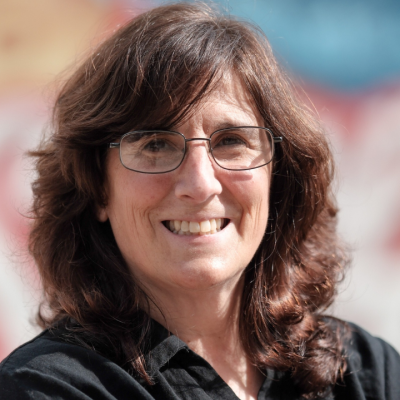 Smiling person with shoulder-length dark brown curly hair and rectangular glasses, wearing a dark collared top