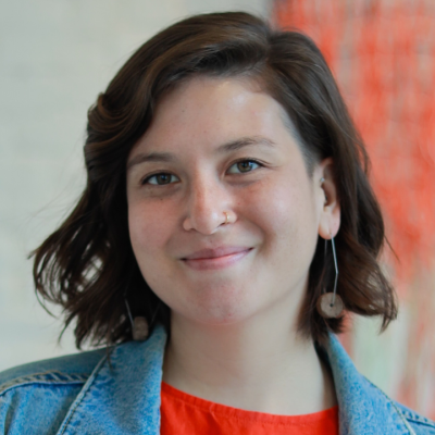 Portrait of a smiling person with shoulder-length dark hair wearing a blue denim jacket and an orange top