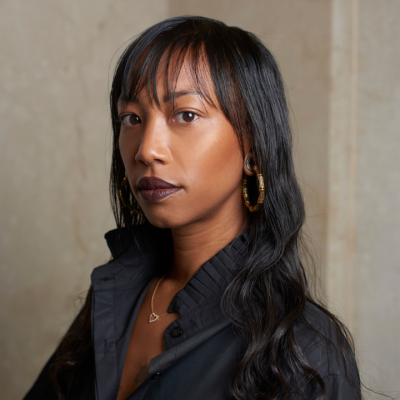 Woman with long dark hair and black shirt standing against a beige background.