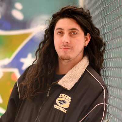 A person with long hair leans against a chain link fence outdoors.