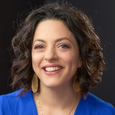 Smiling person with shoulder-length wavy dark hair wearing a bright blue top, gold hoop earrings, and a thin gold necklace