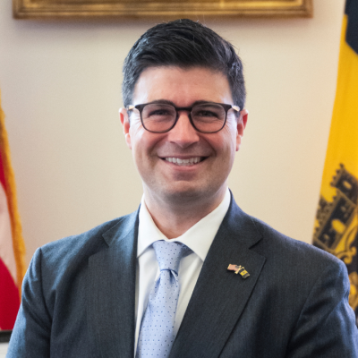 Man in suit stands smiling in office between two flags