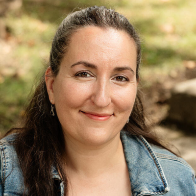 Smiling person with dark hair pulled back, wearing a denim jacket over a dark top