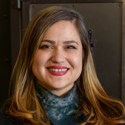 Smiling person with straight shoulder-length brown hair, wearing a dark blazer and a blue patterned scarf