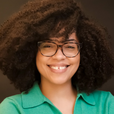Smiling person with curly dark hair and rectangular glasses, wearing a teal green collared top
