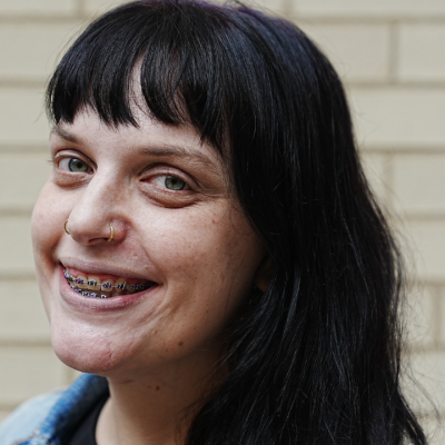 Close-up portrait of a person with long dark hair with nose rings and braces, wearing a jean jacket over a black shirt