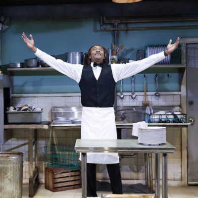 A man stands in a restaurant kitchen with arms raised dramatically.