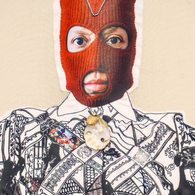 A person wearing a red balaclava exposing their eyes and mouth, dressed in a patterned black and white shirt with a large decorative brooch at the collar, standing against a light-colored background.