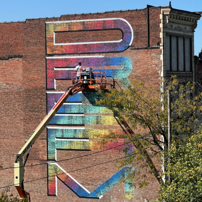Two people in a lift paint a colorful, large-lettered mural on a brick building wall.