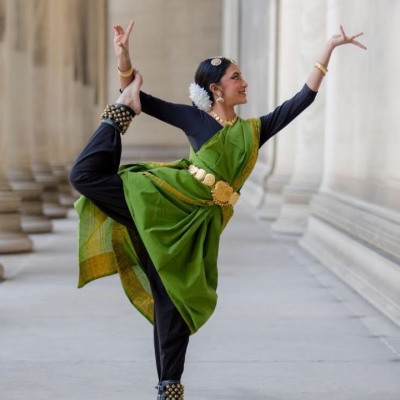 A woman in green Indian traditional attire poses in a classical dance stance between tall columns.