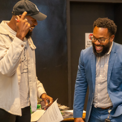 Two men smiling and laughing together indoors; one wears a cap and light jacket, and the other wears glasses and a blue blazer. They appear to be having an animated, friendly conversation.