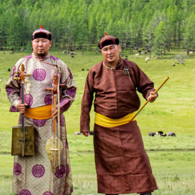 Three men in traditional Mongolian clothing stand on a grassy field with trees and grazing animals in the background. One holds a stringed instrument, and all wear hats and bright yellow sashes.