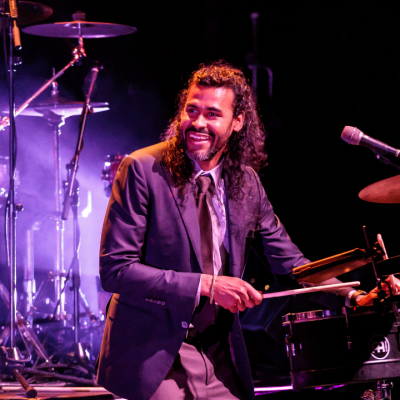 Man in suit playing drums on stage with purple lighting in the background.