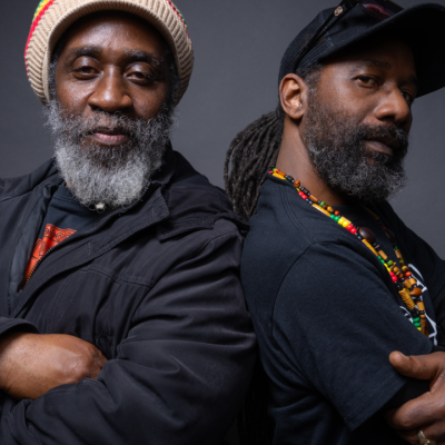 Two men stand back to back with arms crossed, both wearing black shirts. One wears a knit hat and jacket, the other a cap and beaded necklaces. They both have beards and confident expressions against a dark background.