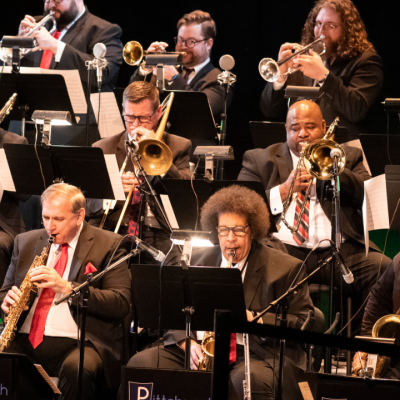 A jazz orchestra performs on stage with various brass and woodwind instruments.