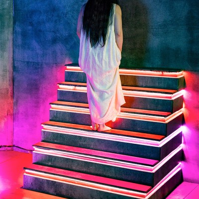 A person in a long white dress with long hair stands on illuminated steps with glowing pink and purple lights, facing a dark textured wall in a vibrant, modern setting.