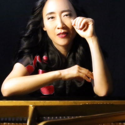 Woman sitting at a grand piano, looking forward with hand raised near her face.
