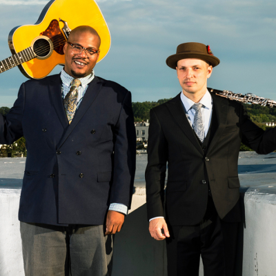 Two men in suits stand outside; one holds a guitar, the other holds a clarinet.