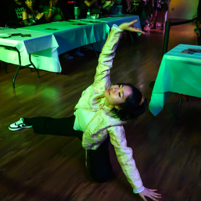 A person poses on a wooden floor in front of tables at an indoor event.