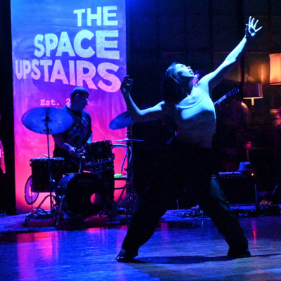A dancer performs on stage with a band under blue and purple lighting.