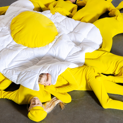 People in yellow bodysuits form the shape of a fried egg on a gray floor.