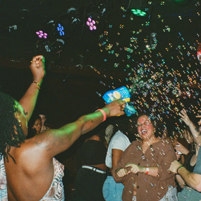 A lively crowd dances as a dancer sprays bubbles at a nightclub with colorful lights.